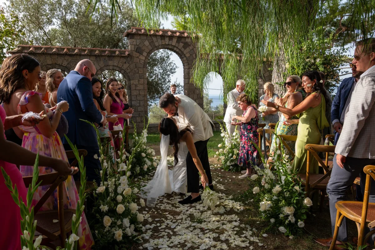 amalfi coast wedding ceremony engaging events ali capo santa fortunata 1 - An Effortless Amalfi Coast Celebration Bride and groom kiss during outdoor wedding ceremony at Capo Santa Fortunata in Sorrento Italy planned by Engaging Events by Ali with white floral arrangements and stone archway backdrop